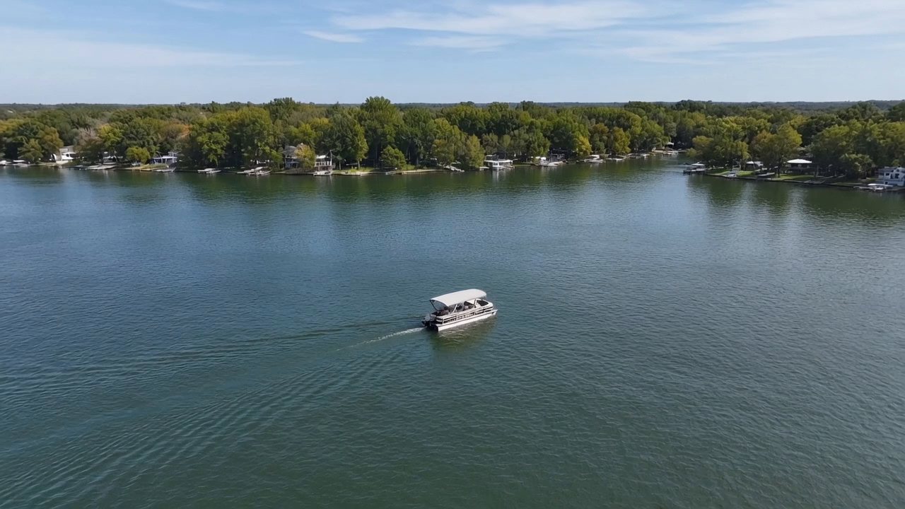 An aerial view of a pontoon boat crossing a wide lake, a wooded shore with houses and docks behind it